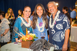 A group of three stand smiling at an event table together, each wearing a lei.