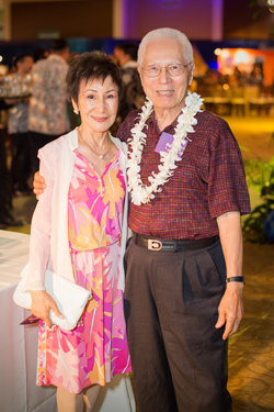 Two people pose for a photograph at an event, each wearing a lei of white flowers.