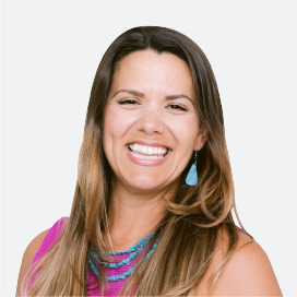 A dark-haired woman smiles kindly, wearing a bright pink shirt and turquoise jewelry.