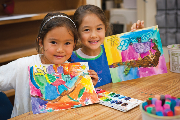 Two young girls smiling at the camera while holding their artwork