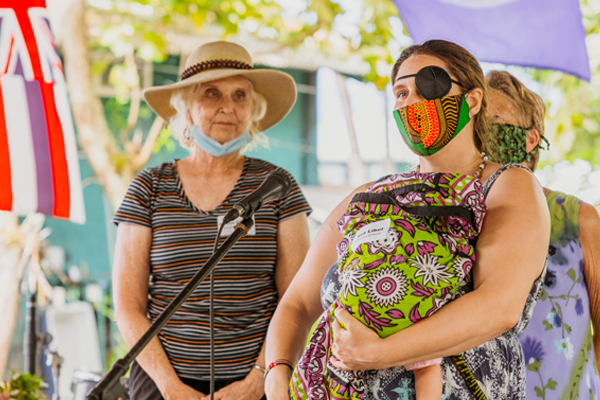 Women gather at an event, each wearing face masks. One holds a baby and wears a black eyepatch.