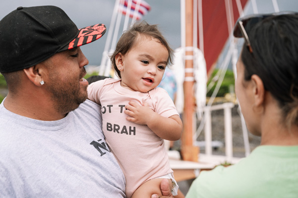 A man holds a young baby near a sailboat.